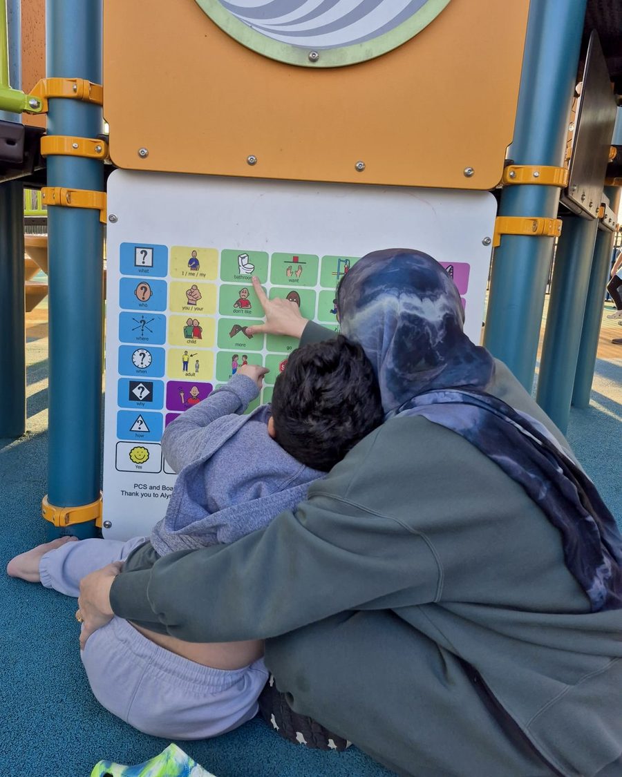 Nazia sitting on the ground with her son at a playground communication board, both seen from behind